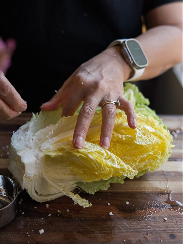 A person wearing a watch and ring is preparing Napa cabbage on a wooden cutting board. Their hands are gently separating the leaves.